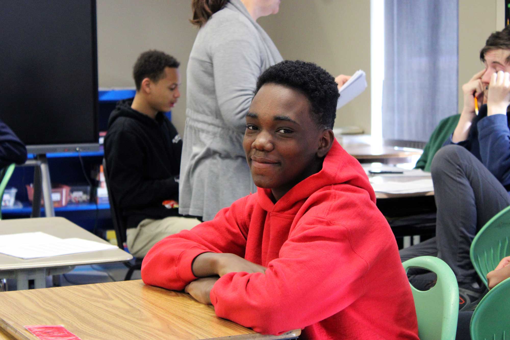 Student in a red hoodie smiling at a desk in a classroom.
