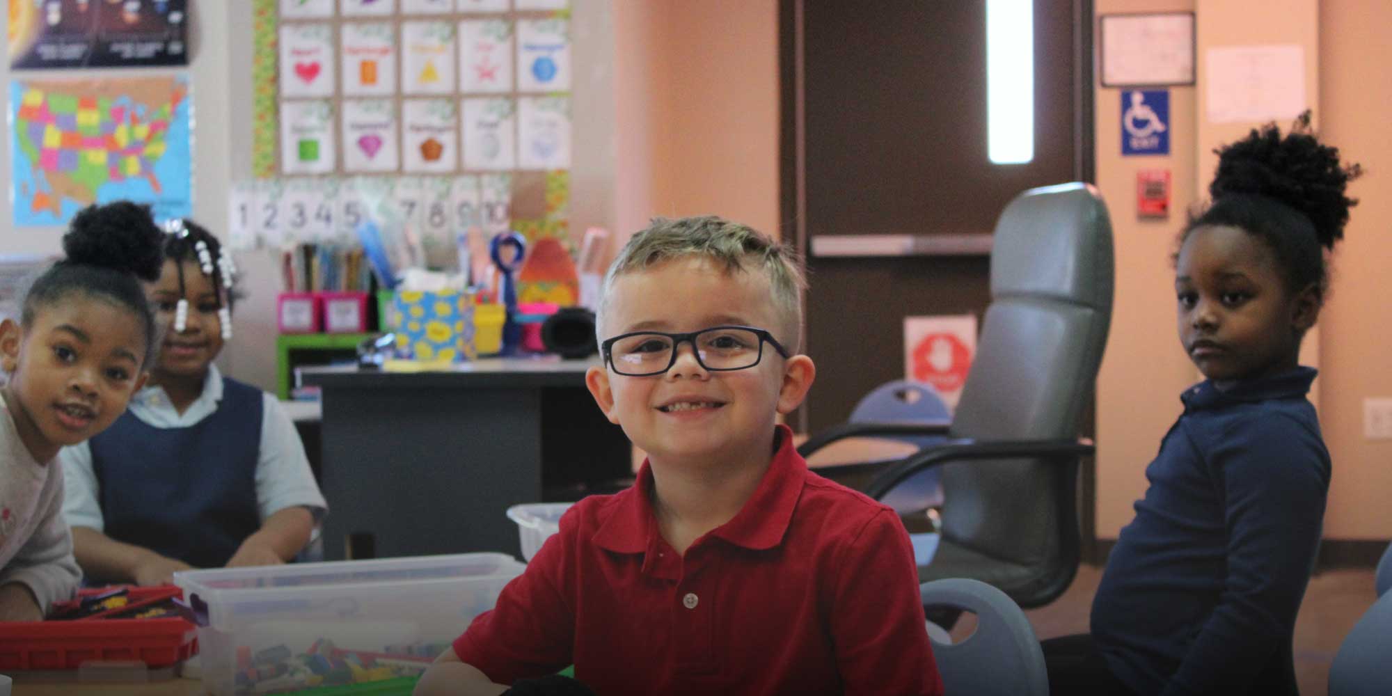Smiling student in glasses and a red shirt with classmates in a classroom.